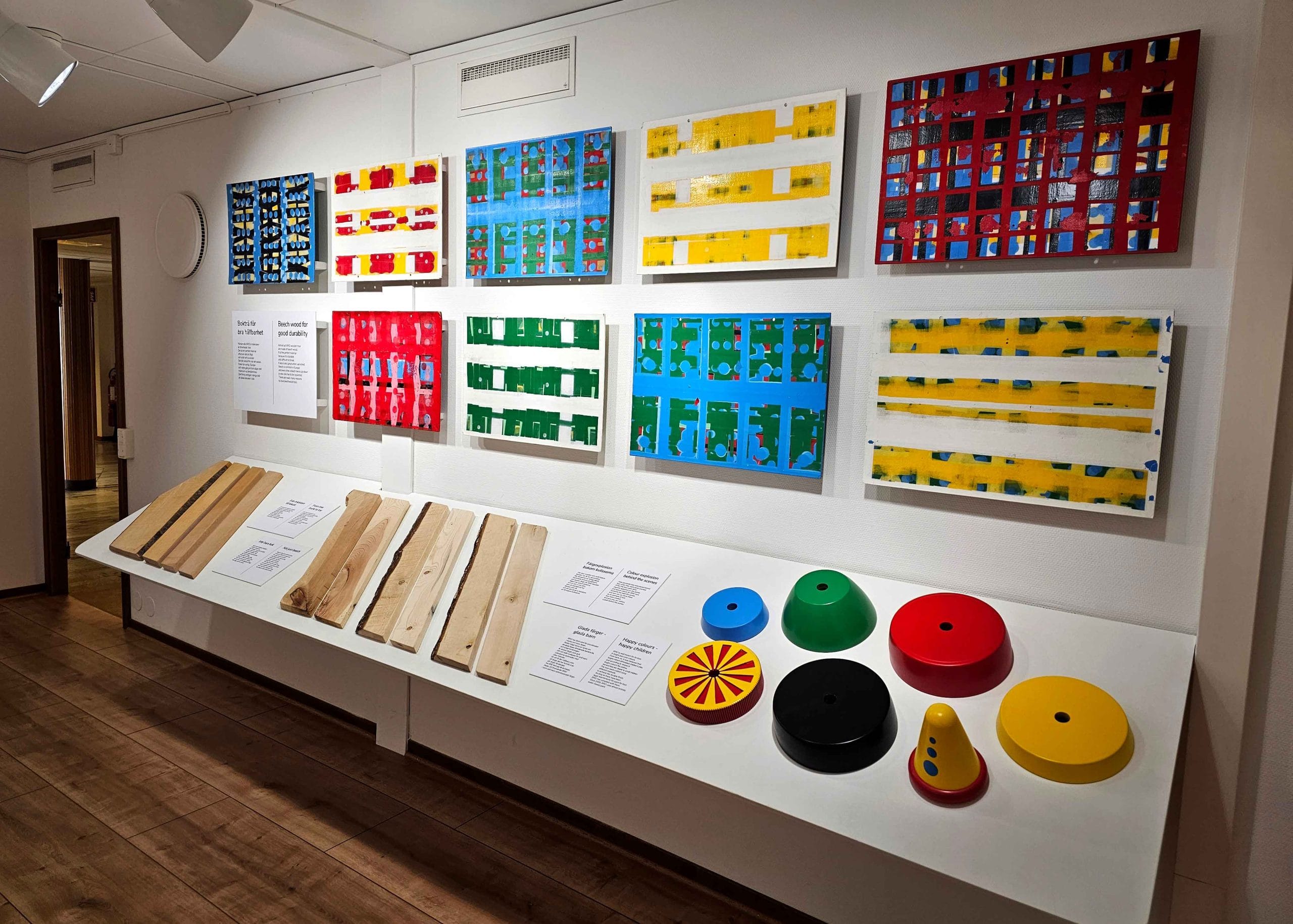 2023-2105-besoka-osby-detaljer-foto-jeanette-junge Vägg med färgrika mönsterbilder ovanför display med träslag och geometriska former i starka färger i utställningsmiljö