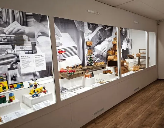 Utställning på Lekoseum i Osby med BRIO-leksaker och hantverk i montrar längs en museivägg