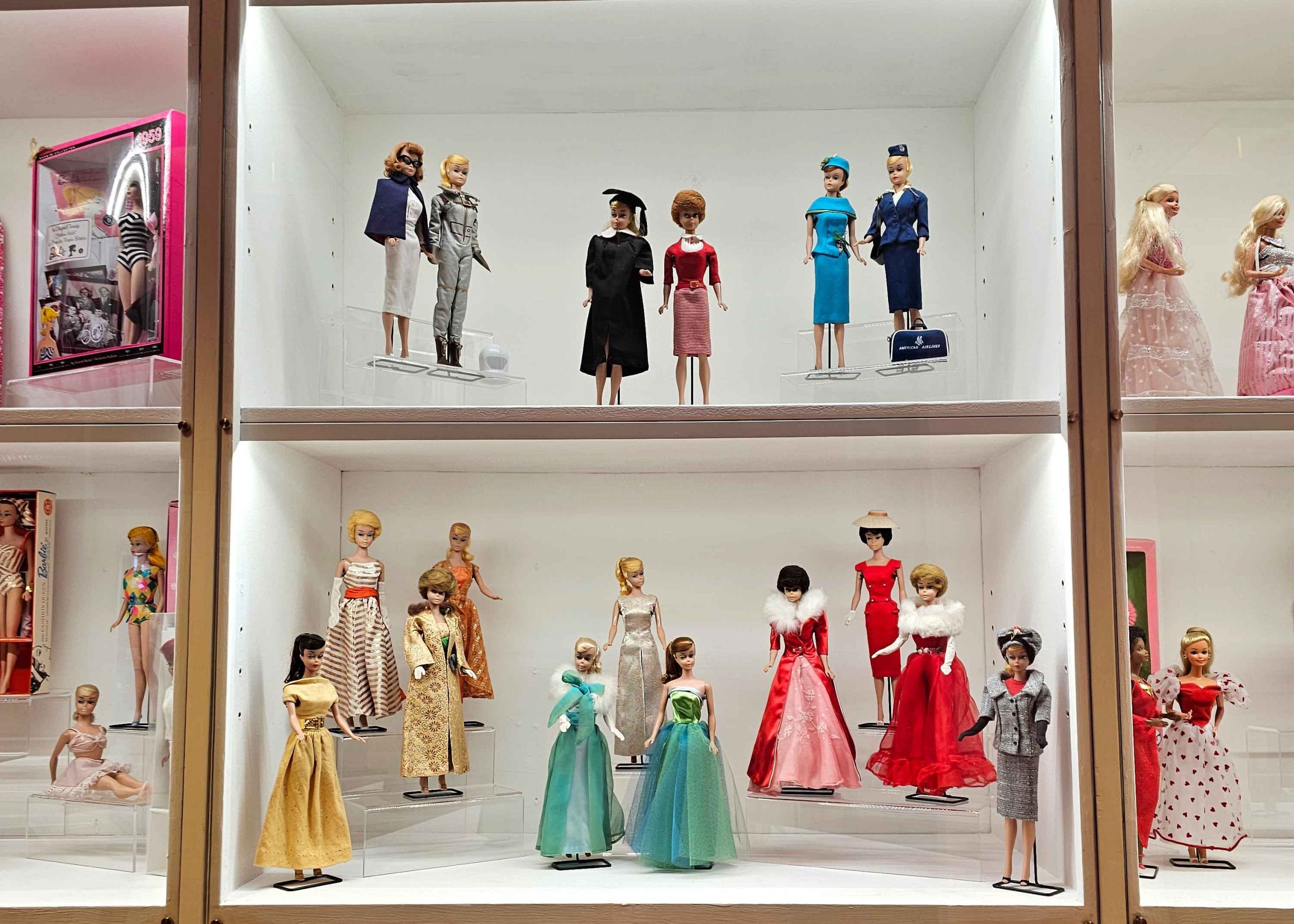 2023-2105-besoka-osby-monter-barbie-foto-jeanette-junge Monter med Barbie-dockor i olika klänningar och yrkeskläder, uppställda i två hyllplan i museiutställning.
