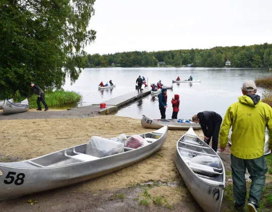 Kanoter vid strand och brygga där människor förbereder paddling i sjö med skog runt Immelnområdet