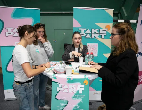 Fyra unga kvinnor står runt ett bor och pratar, runt om finns en massa Take Part-merch