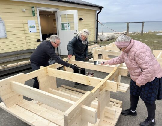 Personer målar och monterar trämöbler utomhus vid kustnära byggnad i Vitemölla som del av utvecklingen av Udden.