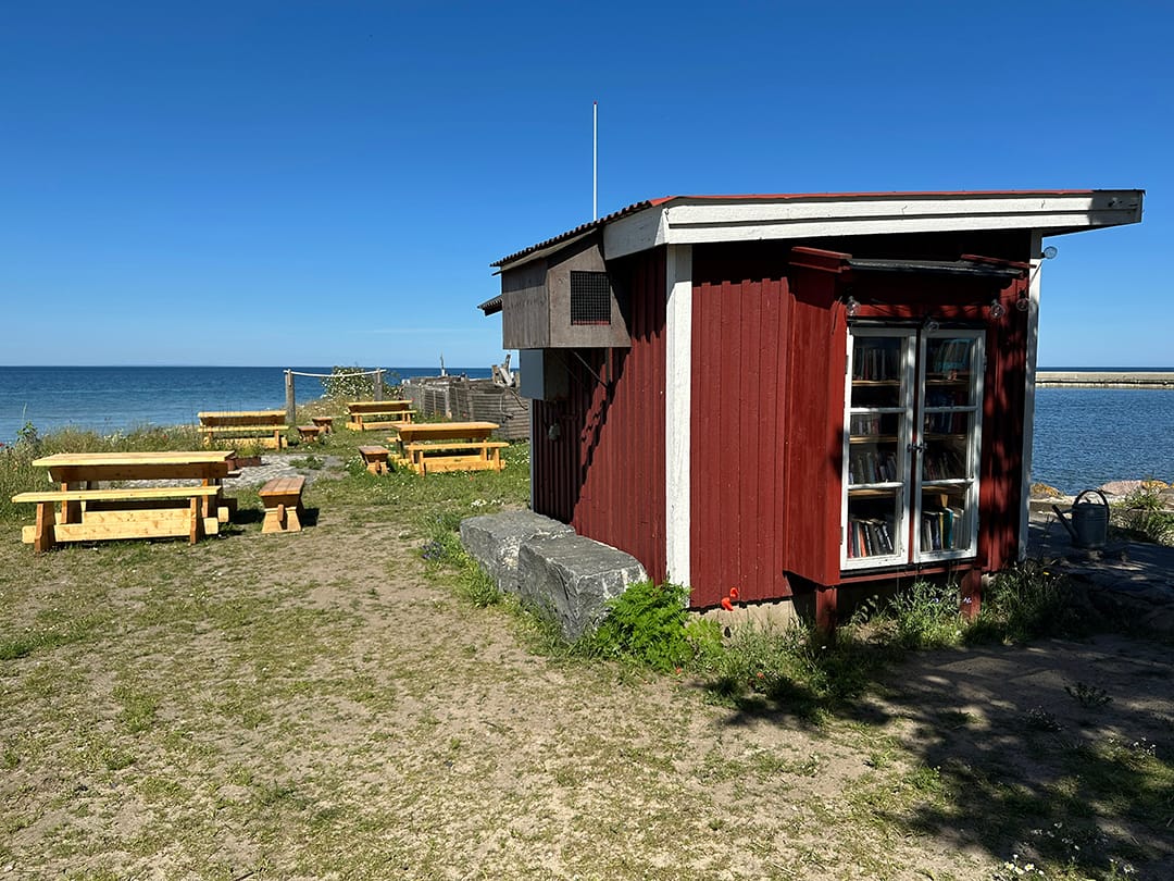 Röd träbod vid havet med ett litet bokskåp i glasdörrar. Runt boden finns picknickbord, gräs, blommor och utsikt över vattnet.