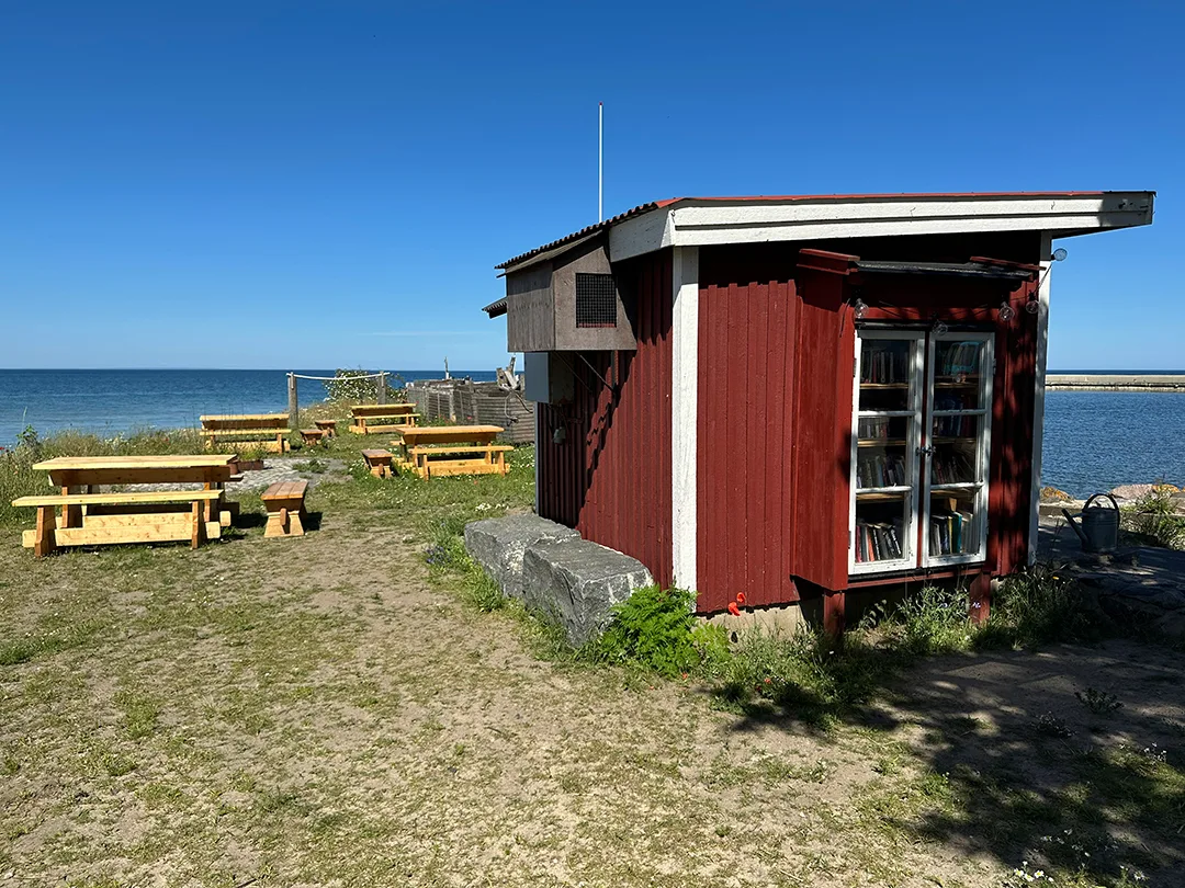 Röd träbod vid havet med ett litet bokskåp i glasdörrar. Runt boden finns picknickbord, gräs, blommor och utsikt över vattnet.