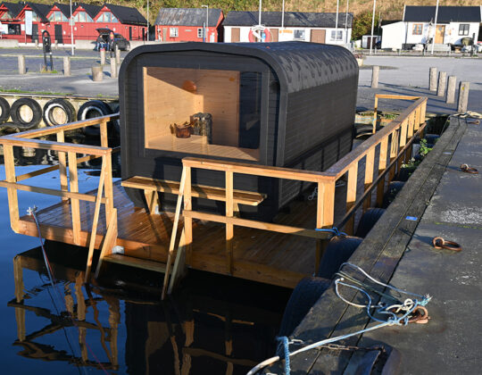 Flytande bastu vid brygga i Kåseberga hamn med trädäck och räcken, förtöjd vid kaj i lugnt vatten