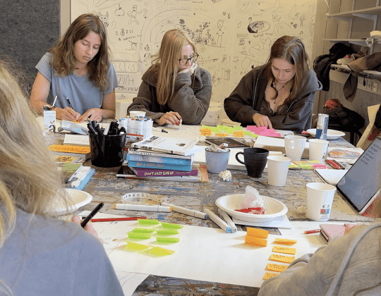 Unga personer sitter runt bord och arbetar kreativt med papper, pennor och post-it-lappar i workshopmiljö