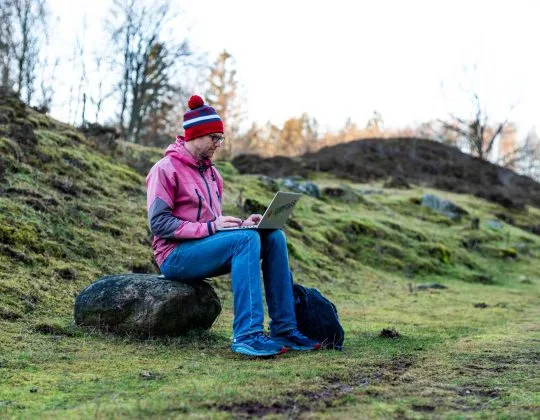 Person sitter utomhus på sten med laptop i naturmiljö och arbetar digitalt i landskap på Österlen