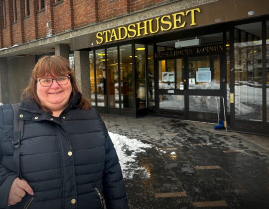 Kvinna i svart jacka står utanför Stadshuset i Hässleholm, med snö på marken och entrén synlig i bakgrunden