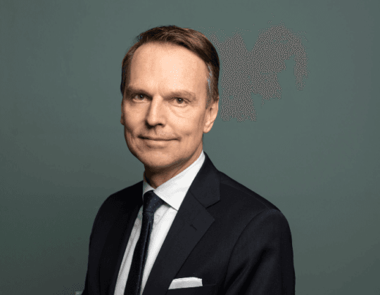 Porträttbild av en man i mörk kostym, vit skjorta och mörk slips. Han står framför en enfärgad grågrön bakgrund och tittar mot kameran med ett lätt leende. Bilden är professionellt tagen med mjukt ljus och neutral ton.