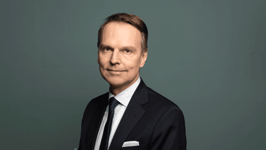 peter-danielsson Porträttbild av en man i mörk kostym, vit skjorta och mörk slips. Han står framför en enfärgad grågrön bakgrund och tittar mot kameran med ett lätt leende. Bilden är professionellt tagen med mjukt ljus och neutral ton.