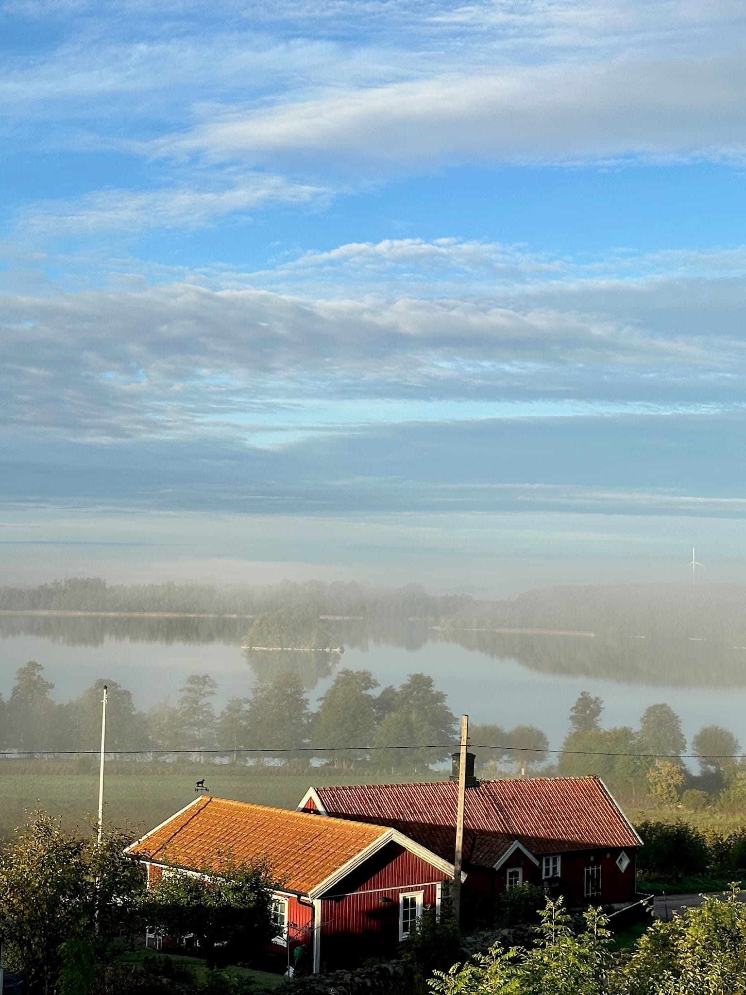 Röd stuga med utsikt över en dimmig sjö med små öar under en blå himmel med moln. Foto: Kerstin Söderlind